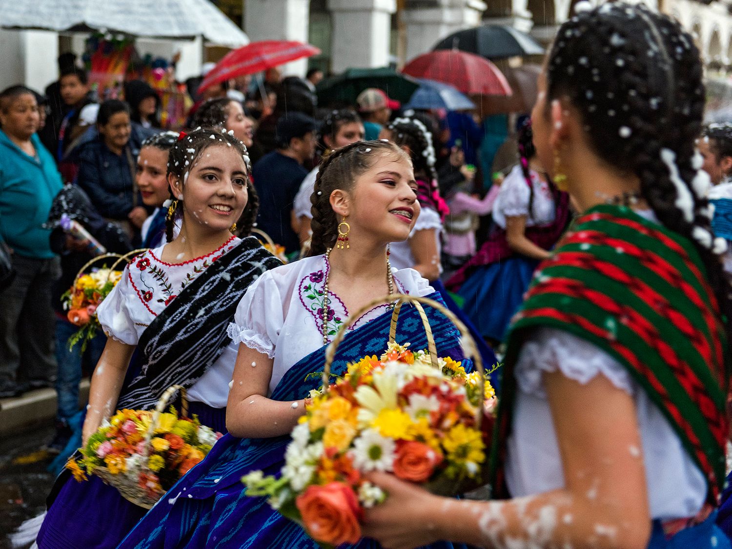 Women in traditional embroidered dresses carry flower baskets during a colorful Mexican cultural parade celebration.