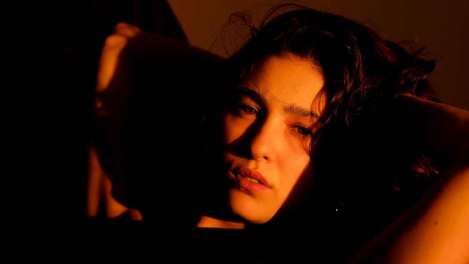Dramatic warm-toned portrait of a young woman with dark wavy hair in moody orange lighting.