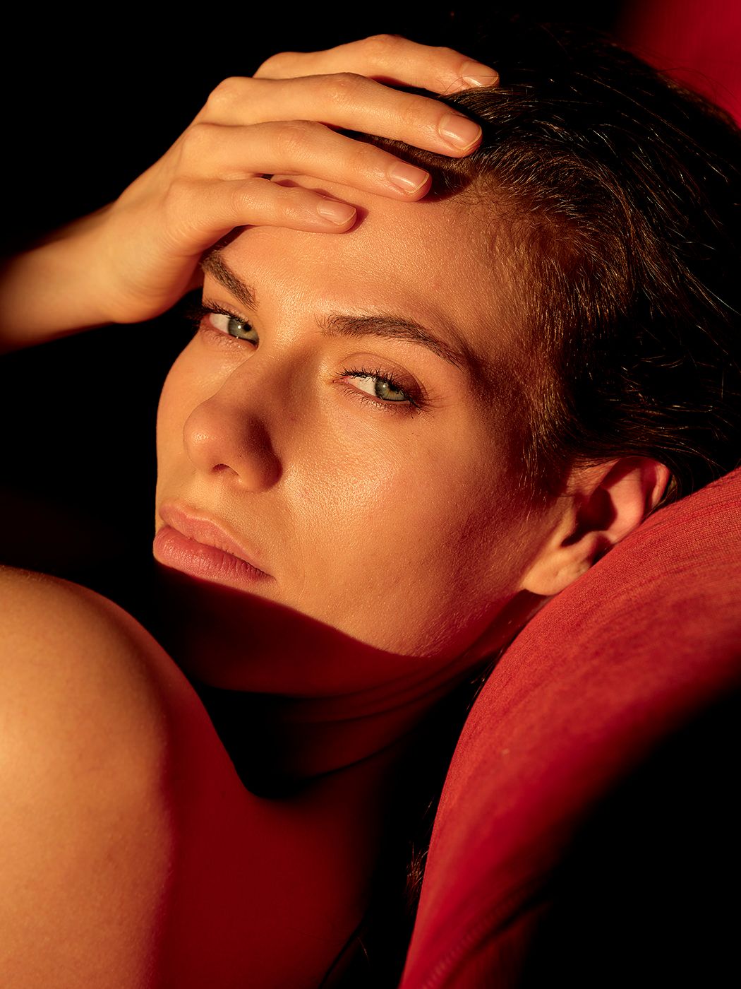 Close-up portrait of a woman resting her hand on her forehead, bathed in warm red and golden light.