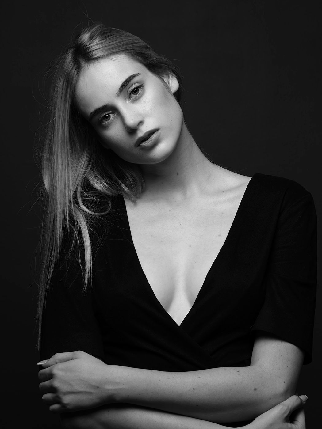 Black and white portrait of a young woman in a deep V-neck black top, with long hair, posing against a dark background.