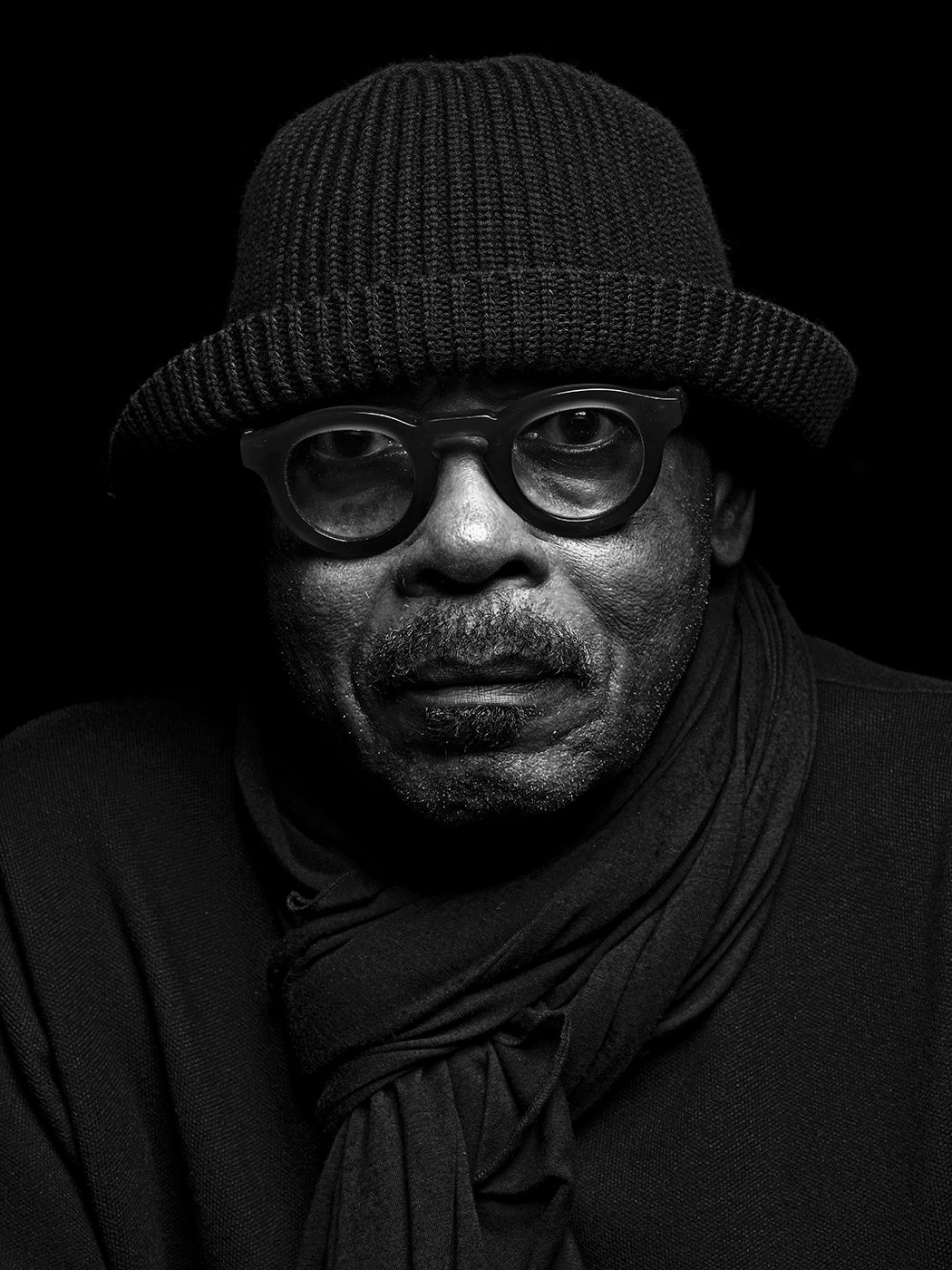 Black and white portrait of an older man wearing round glasses, knit bucket hat, and scarf against dark background. Harvey Mason