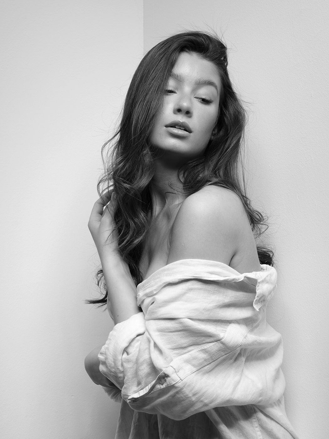 Black and white portrait of a young woman with long wavy hair wearing an off-shoulder white shirt.
