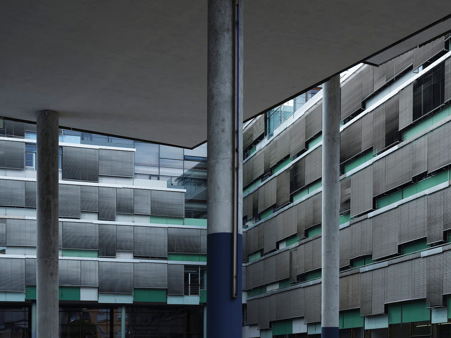 Modern multi-story building facade with horizontal metal louvers, green accents, and concrete columns in an urban setting.