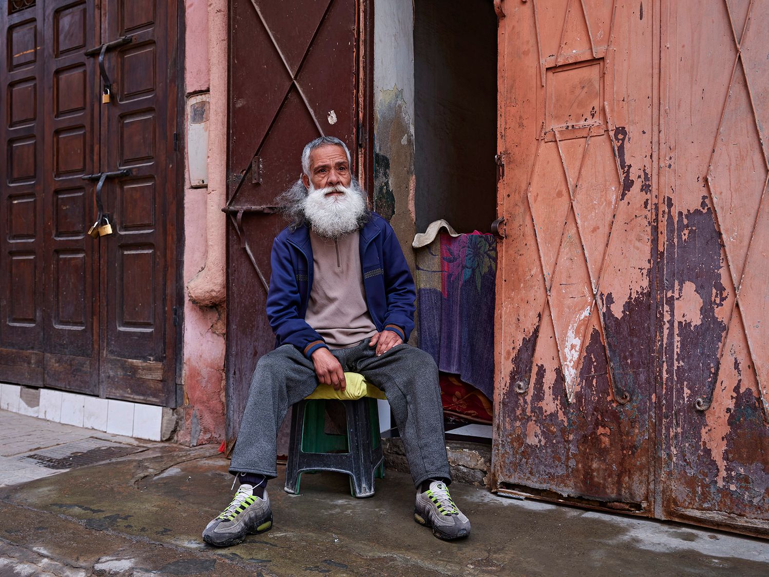 Elderly man with white beard sits on small stool between weathered doors in urban setting.