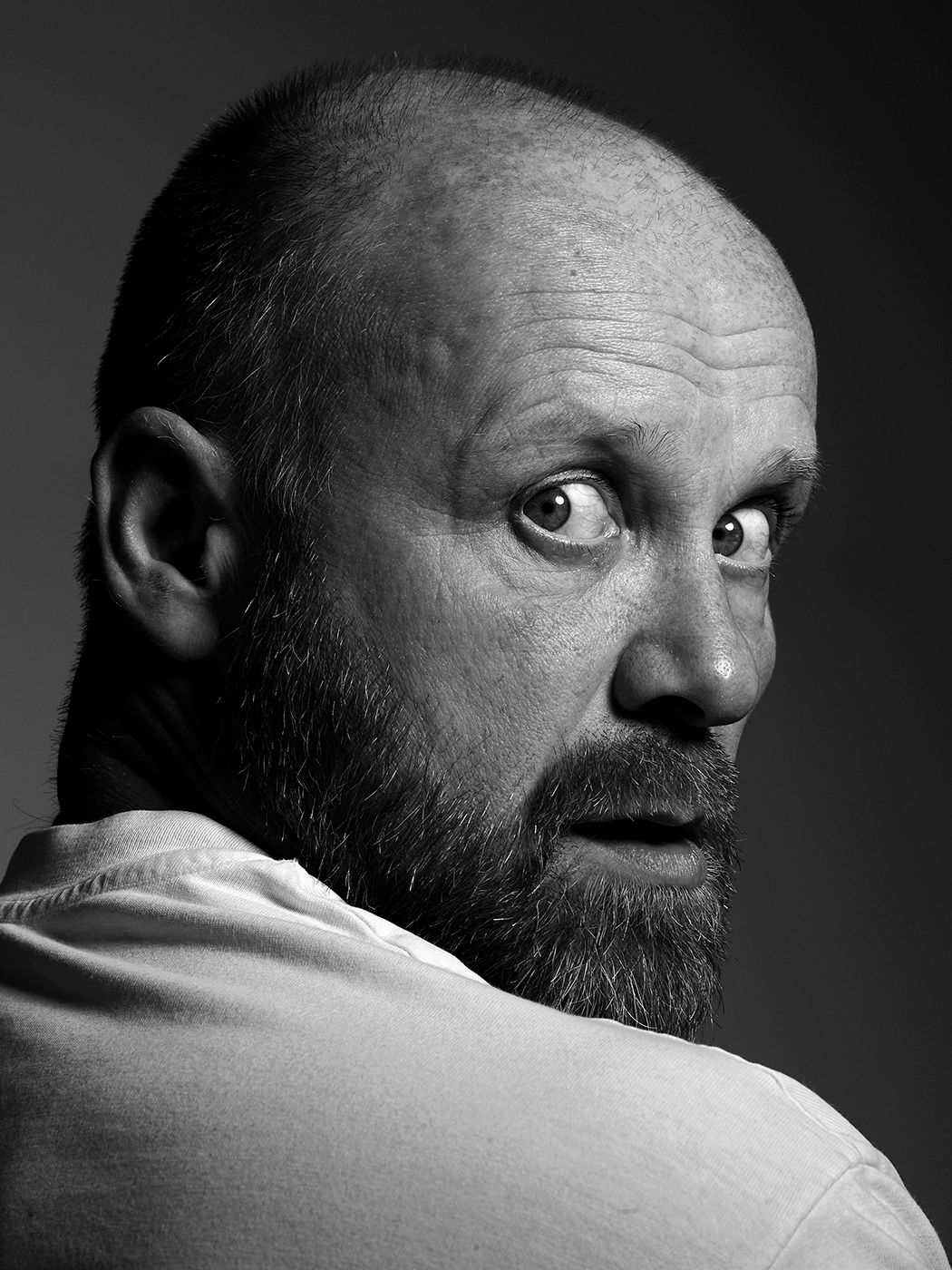 Black and white portrait of a bald bearded man in a white shirt glancing back over his shoulder.