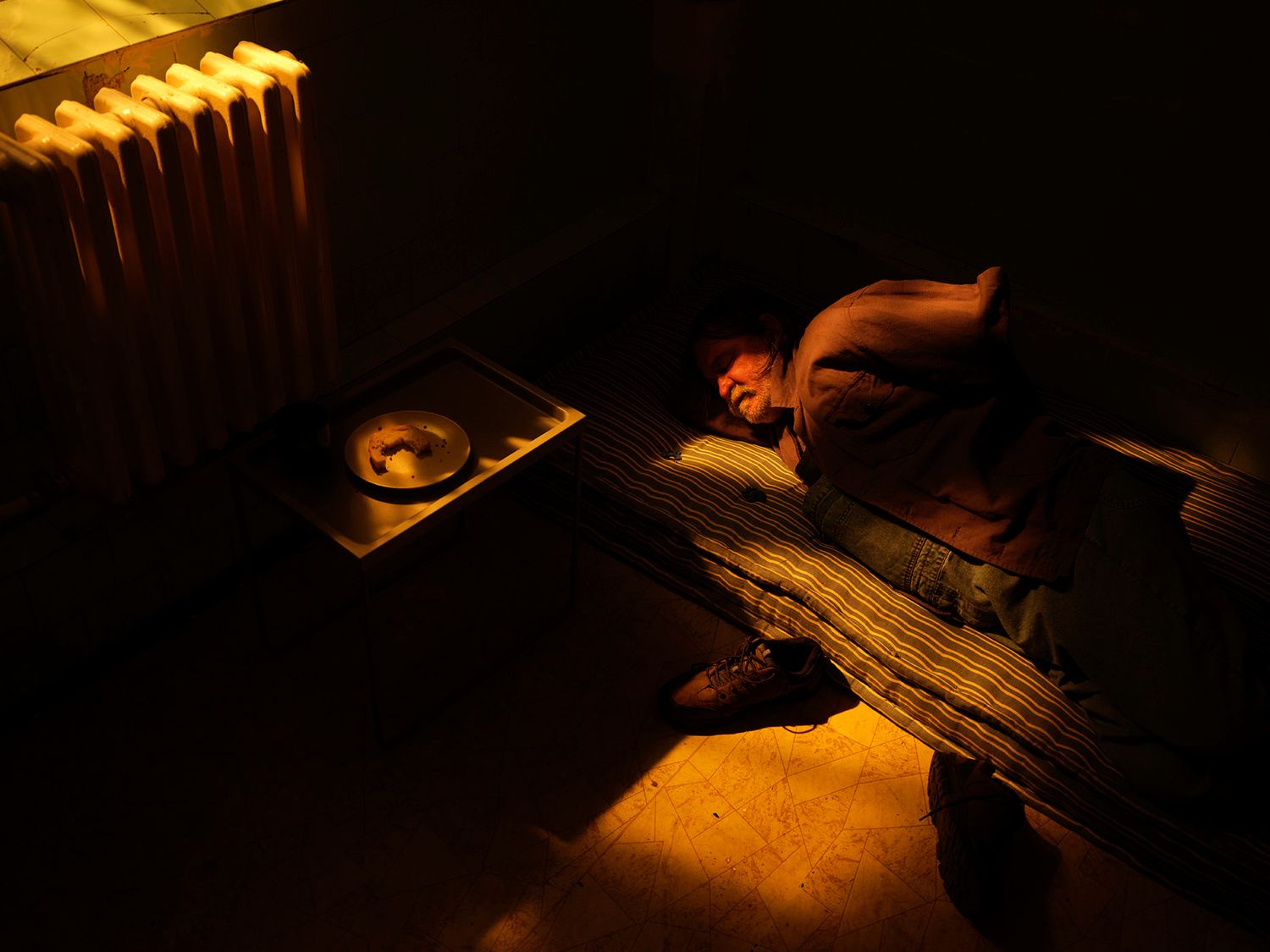 Person sleeping on wooden floor near a radiator in dark moody lighting.