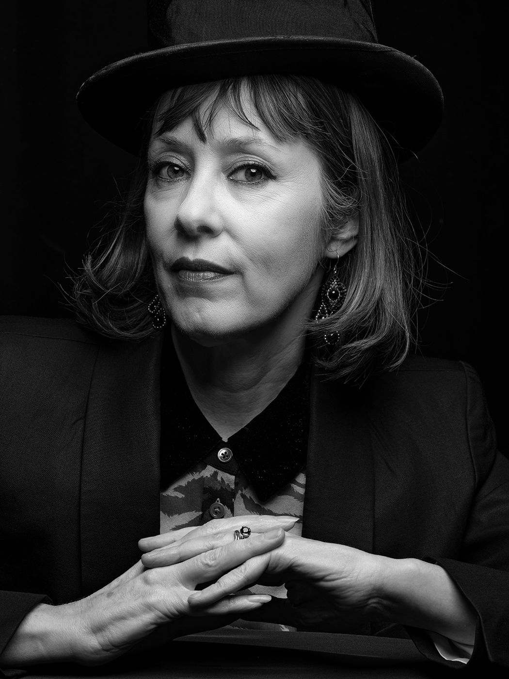 Black and white portrait of a woman wearing a wide-brim hat and blazer, hands clasped, with dramatic lighting. Suzanne Vega, musician.