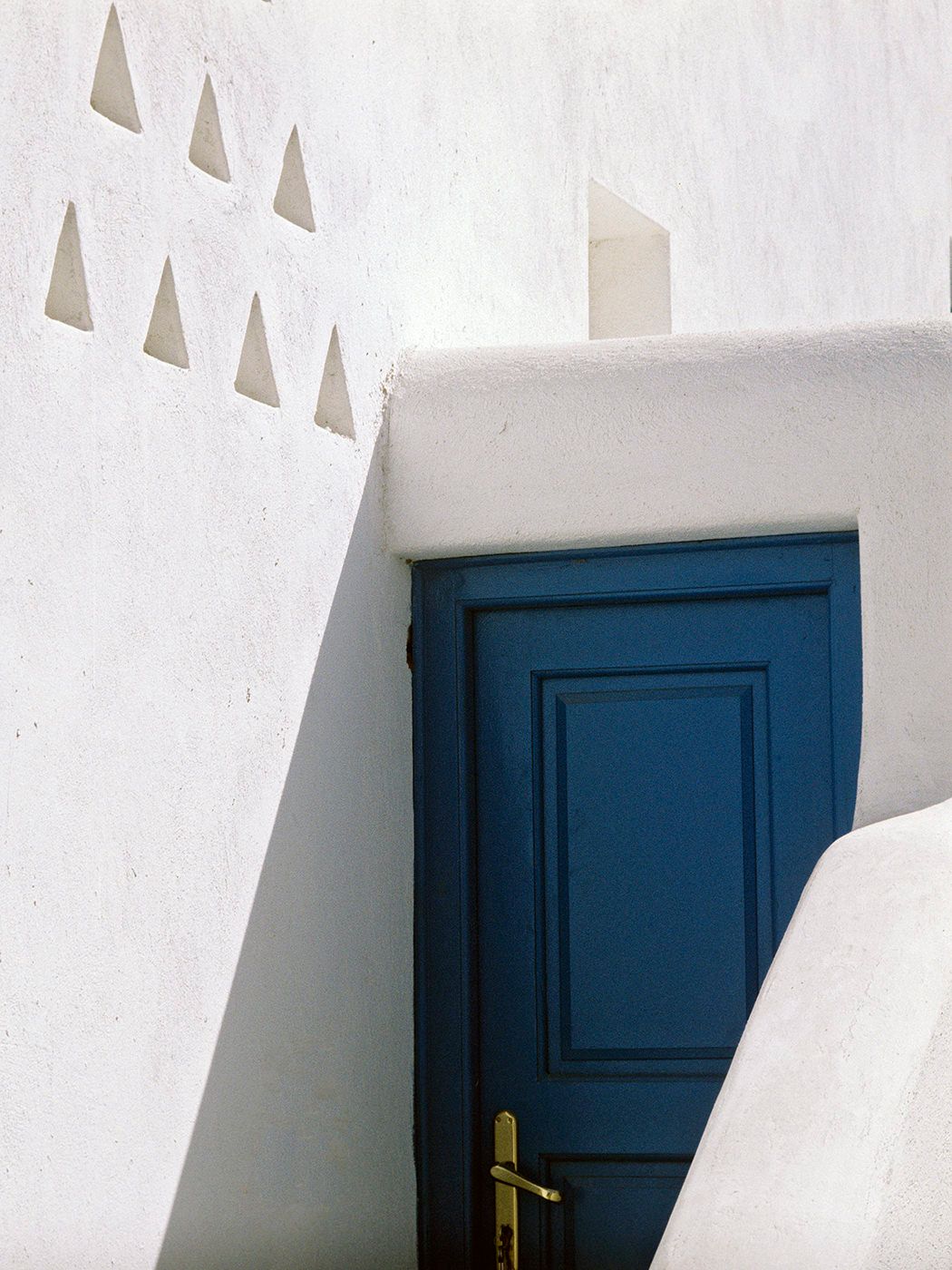 White-washed Greek architecture with a vibrant blue wooden door and gold handle, featuring decorative triangular wall details.