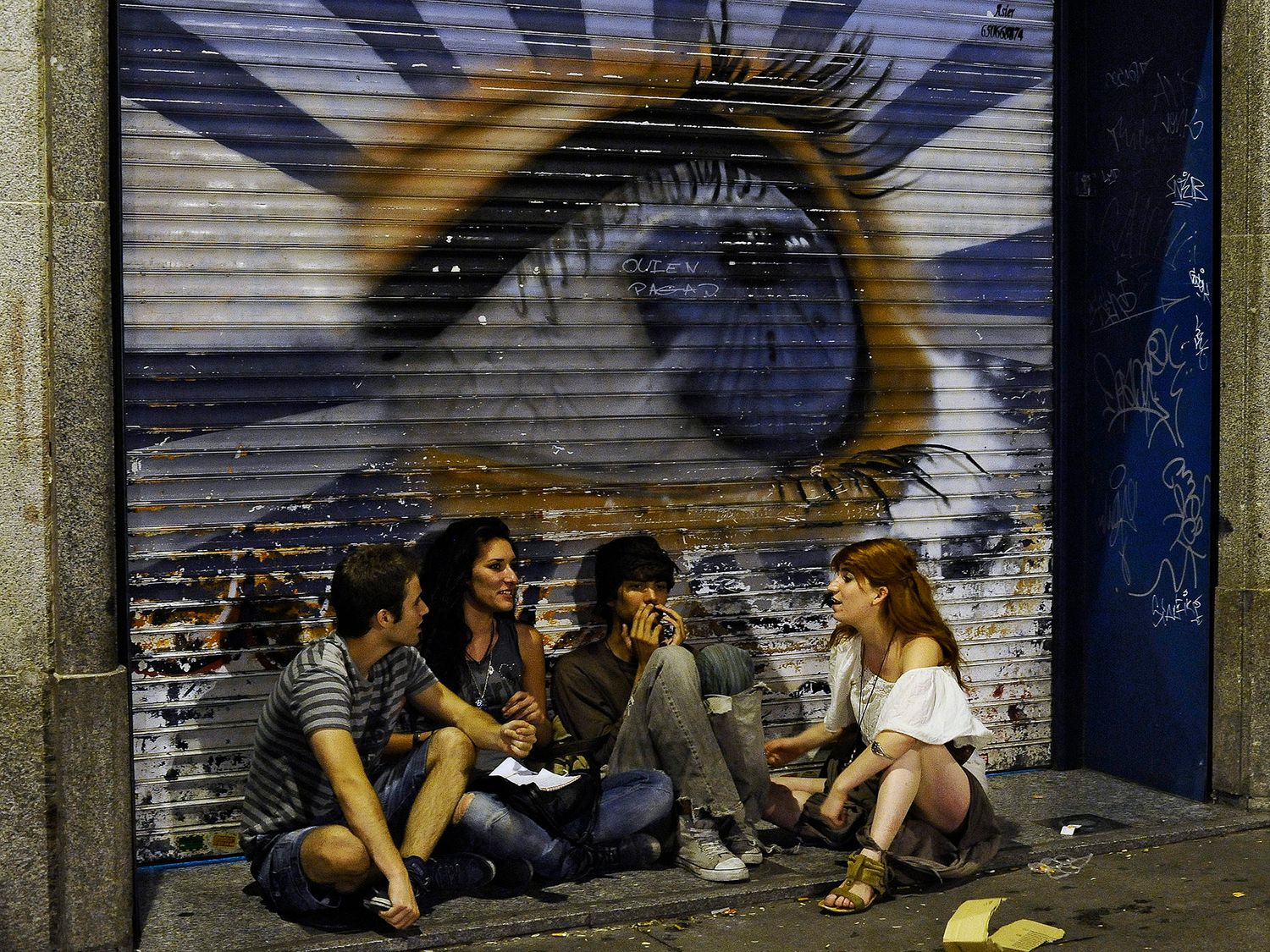 Four young people sitting on a sidewalk in front of a graffiti mural featuring a large eye on a metal shutter.