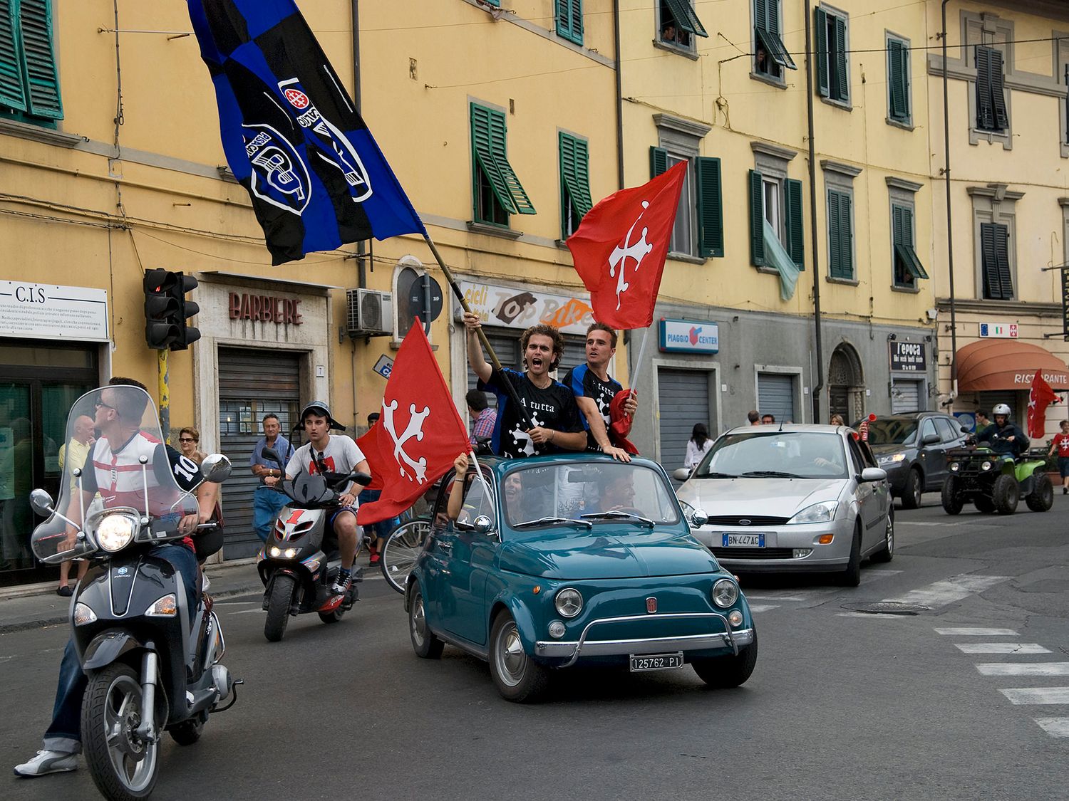 Fans wave red flags from a vintage teal Fiat 500 during a street celebration in an Italian city.