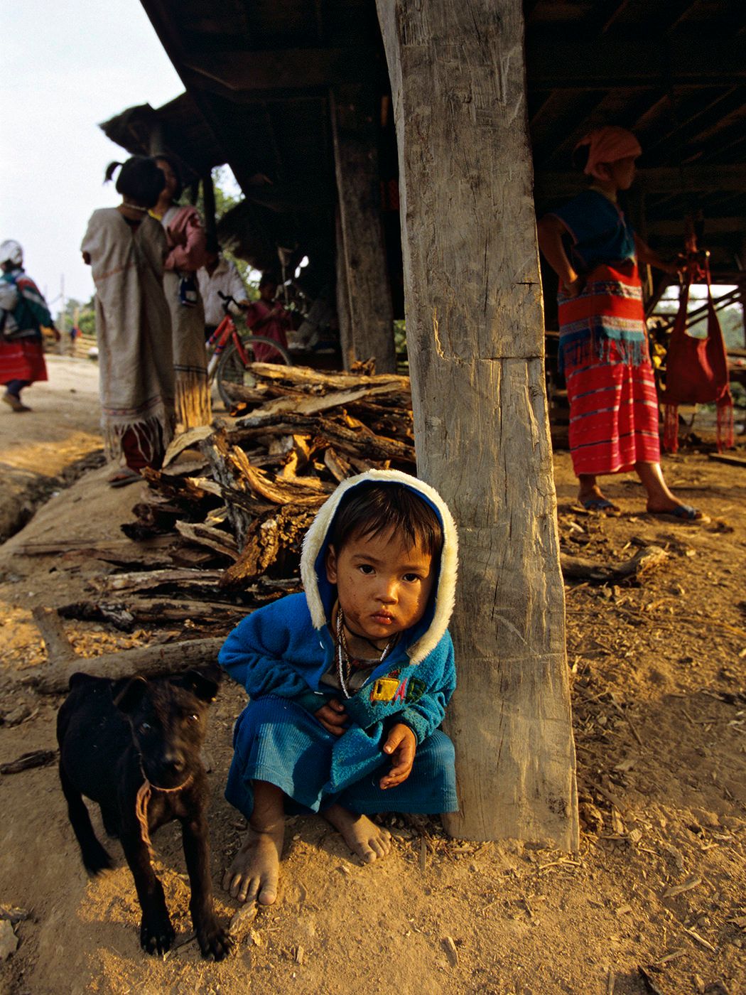 Young child in blue hoodie crouching beside a black goat in a rural village market setting.