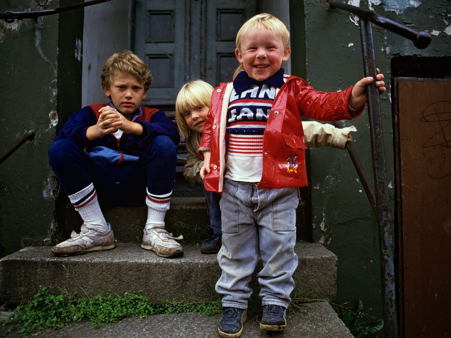 Three young blonde children posing on stone steps near a gate, wearing colorful 1980s clothing.