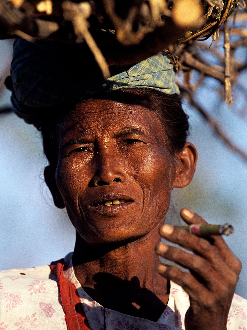 Elderly Southeast Asian woman smoking a cigar outdoors, wearing a plaid headscarf and floral top.