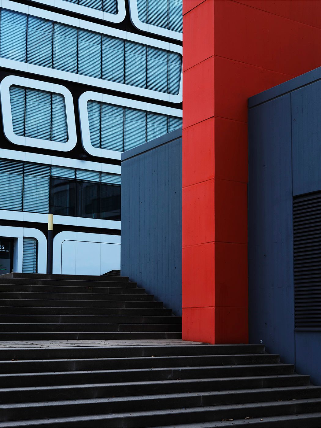 Modern building exterior with bold red column, blue facade, rounded windows, and concrete stairs in urban architecture.