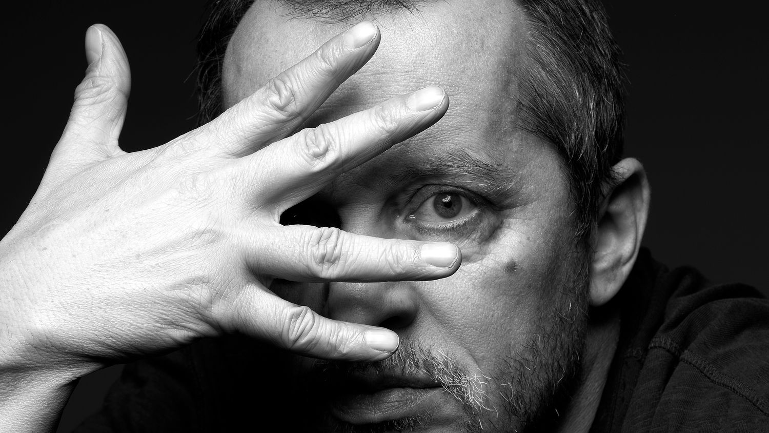 Black and white portrait of a man partially covering his face with his hand, peering through his fingers.