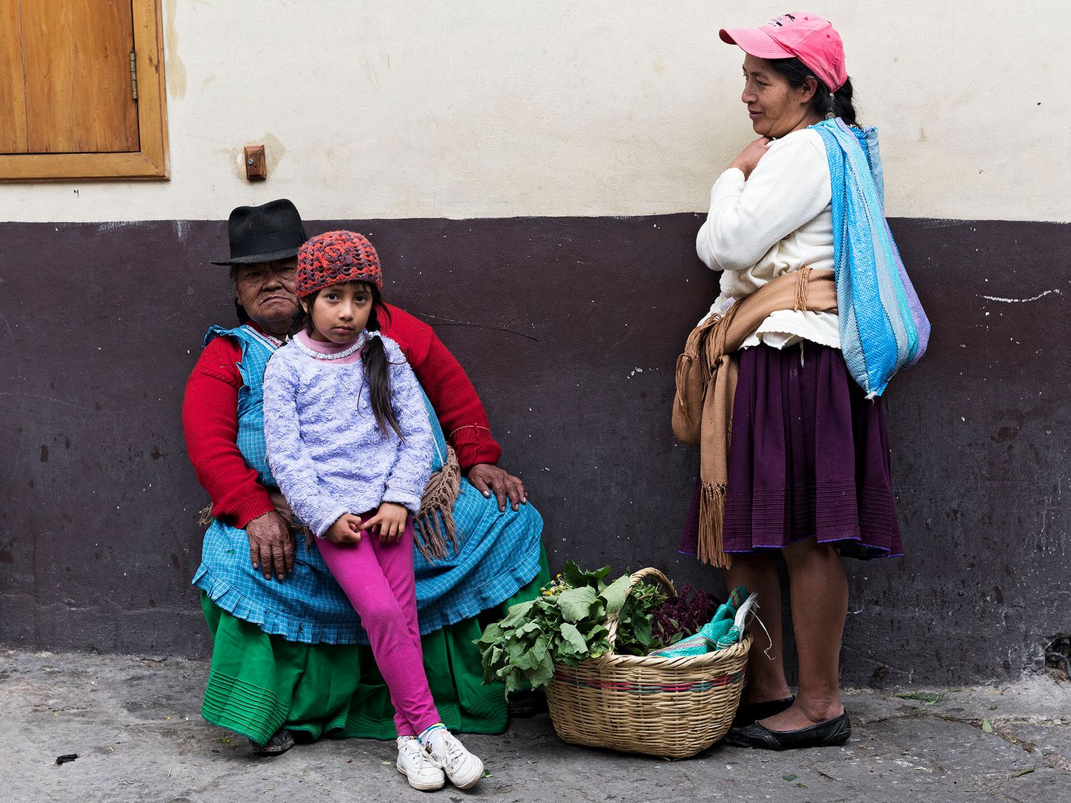Three indigenous women in traditional colorful clothing sit by a basket of vegetables on a street in Ecuador.
