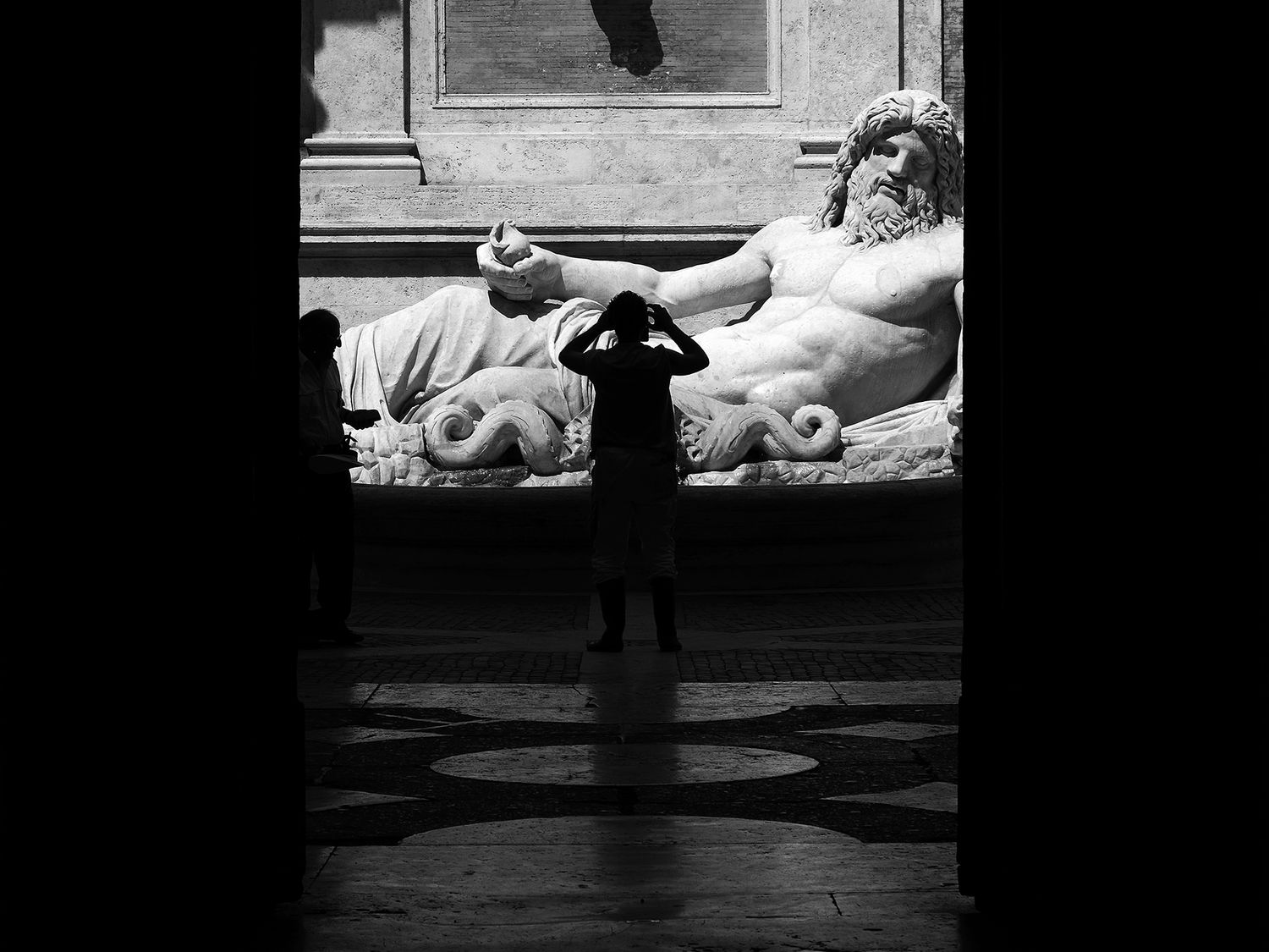 Black and white photo of a child silhouetted before a large reclining Roman sculpture in a museum.