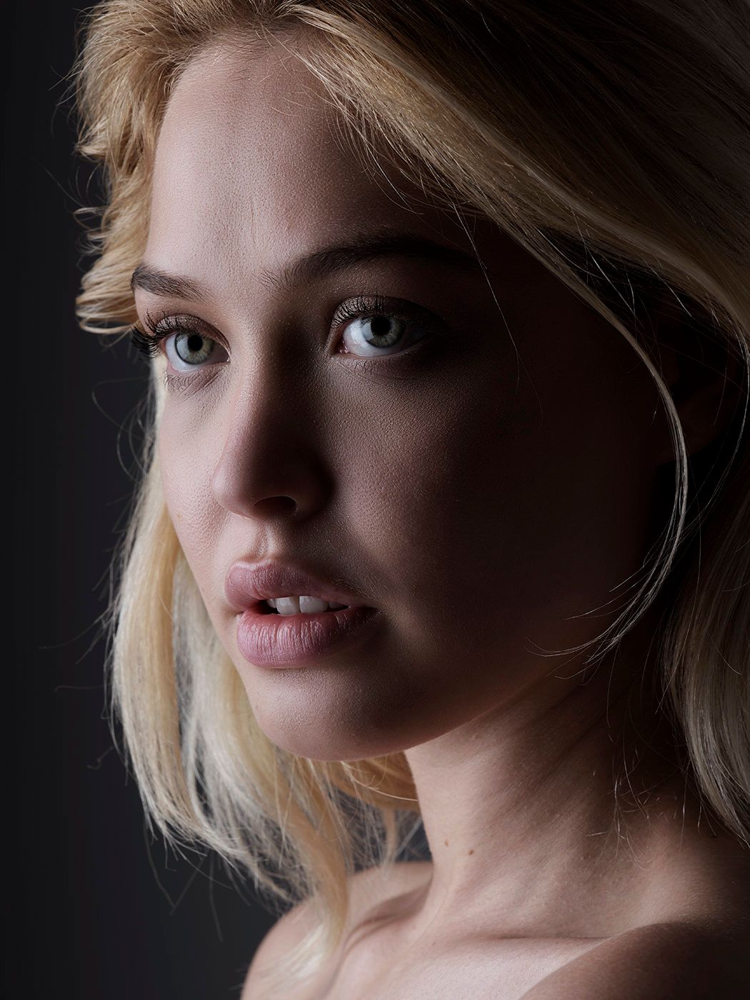 Close-up portrait of a young blonde woman with blue-gray eyes and bare shoulders against a dark background.