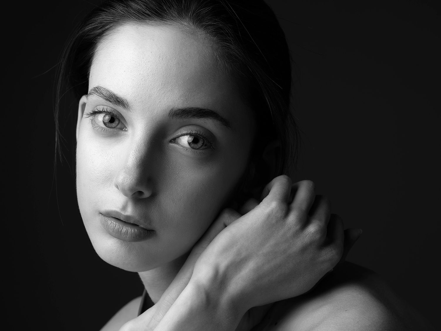 Black and white portrait of a young woman with dark hair, hand on neck, gazing softly at camera.