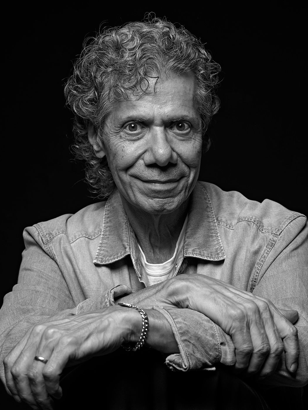 Black and white portrait of an elderly man with curly hair, resting his chin on crossed hands, wearing a denim jacket. Chick Corea