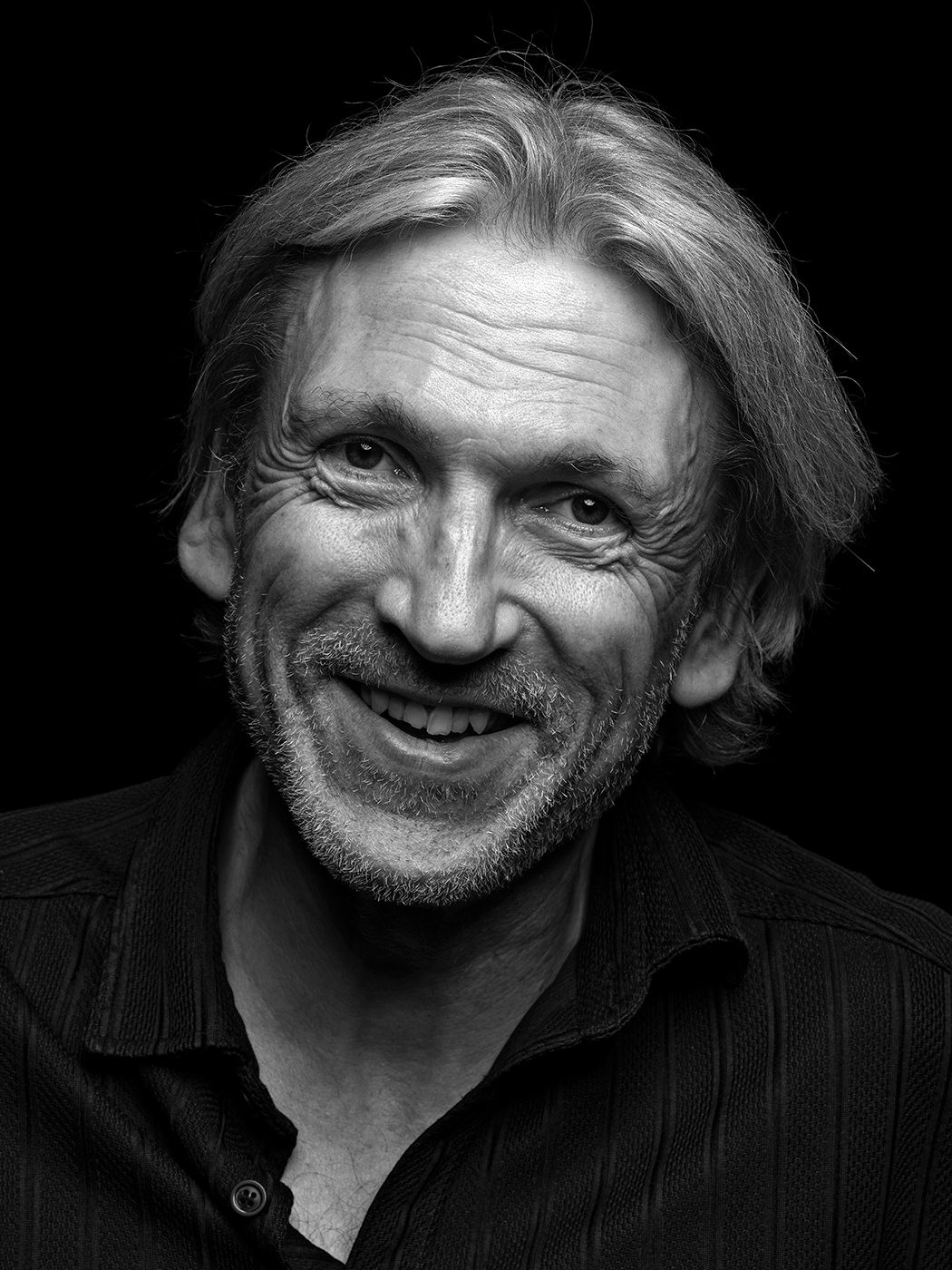 Black and white portrait of a smiling middle-aged man with medium-length hair wearing a dark shirt. Magnus Öström