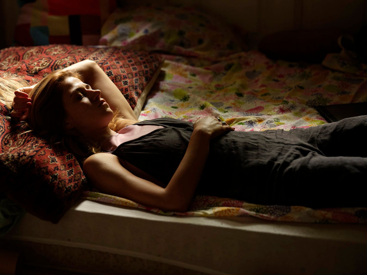 Young woman lying on a bed in dim light, resting with eyes closed.