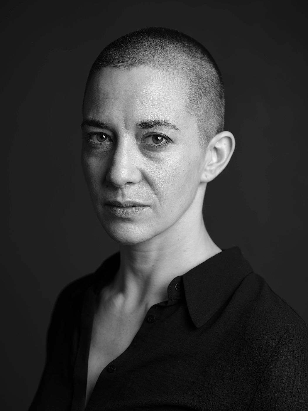 Black and white portrait of a woman with a shaved head wearing a black button-up shirt.