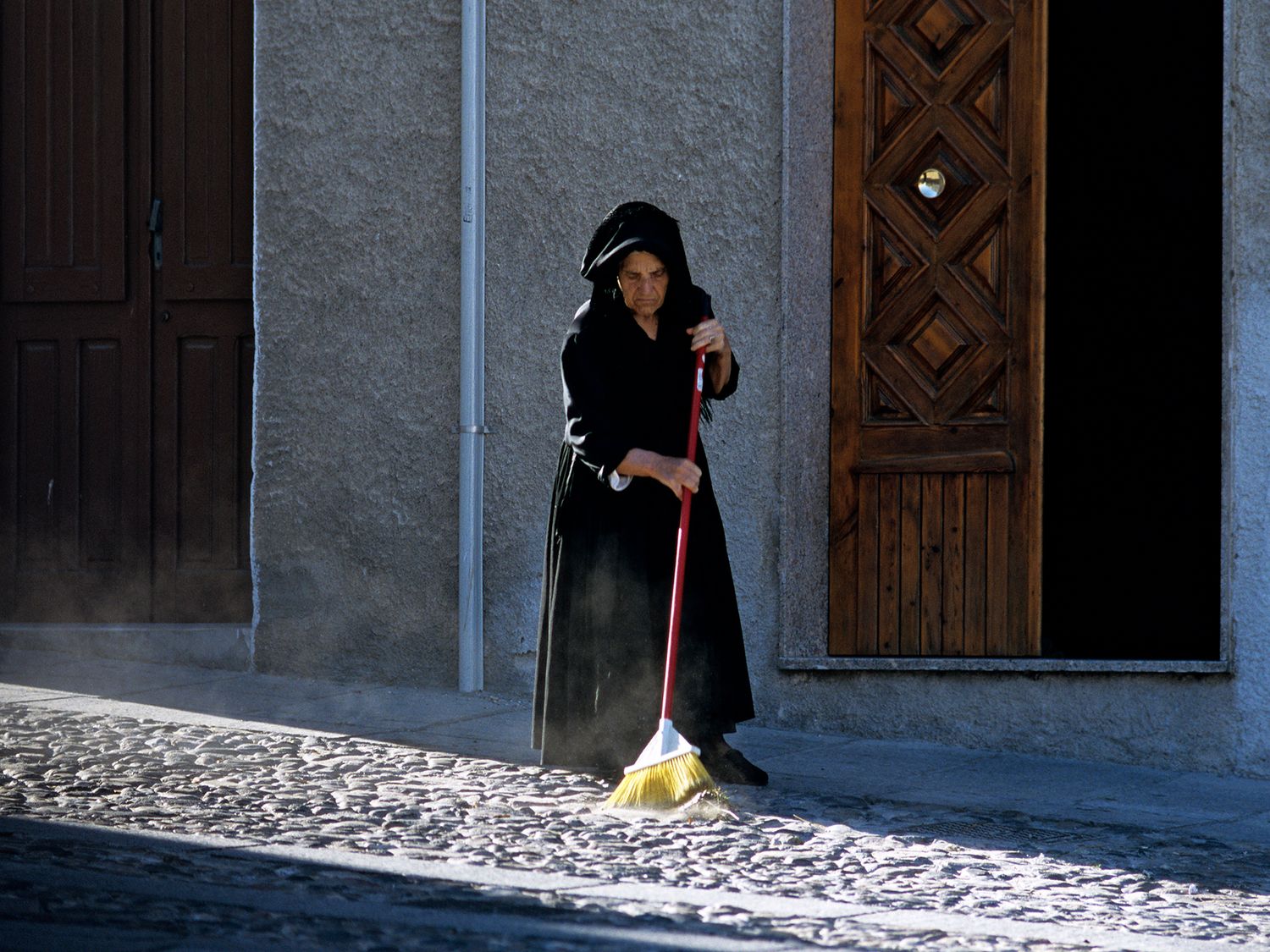 An elderly woman in black dress sweeps cobblestone street with red broom outside historic building doors.