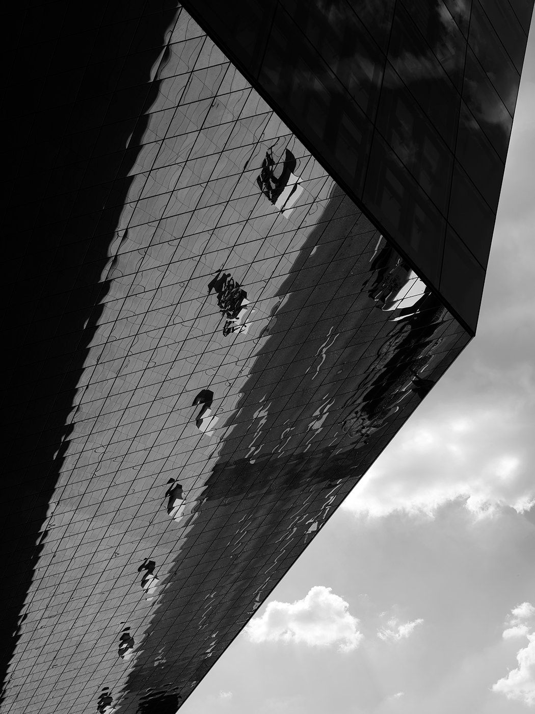 Black and white photo of a glass skyscraper reflecting clouds, shot from a dramatic low angle perspective.