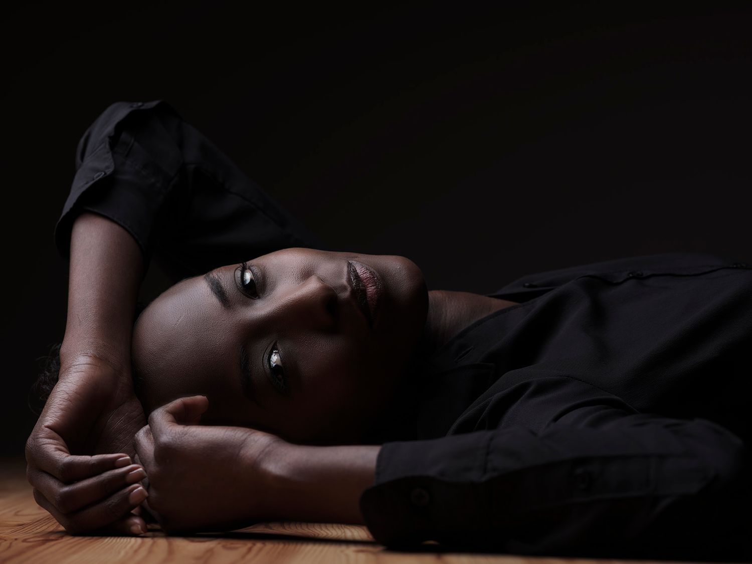 A young Black woman in black clothing lies on a wooden floor, resting her head on her arms in dim light.