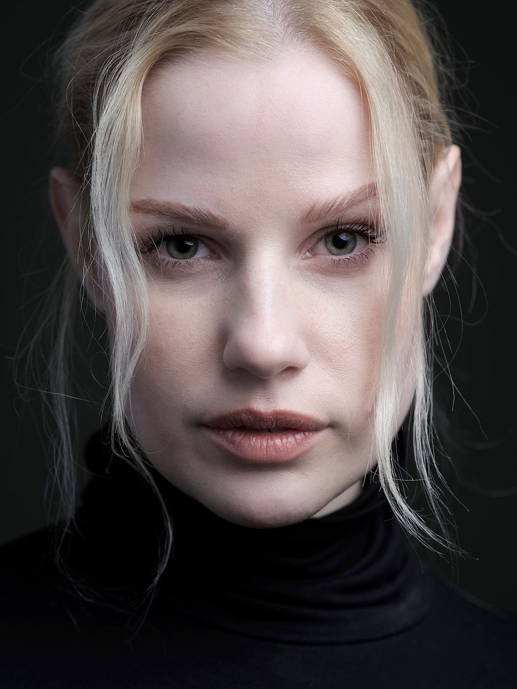 Close-up portrait of a young blonde woman in a black turtleneck against a dark background.