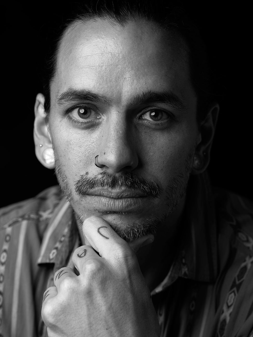 Black and white portrait of a tattooed man with nose ring and mustache, hand on chin, pensive expression. Felix Pastorius