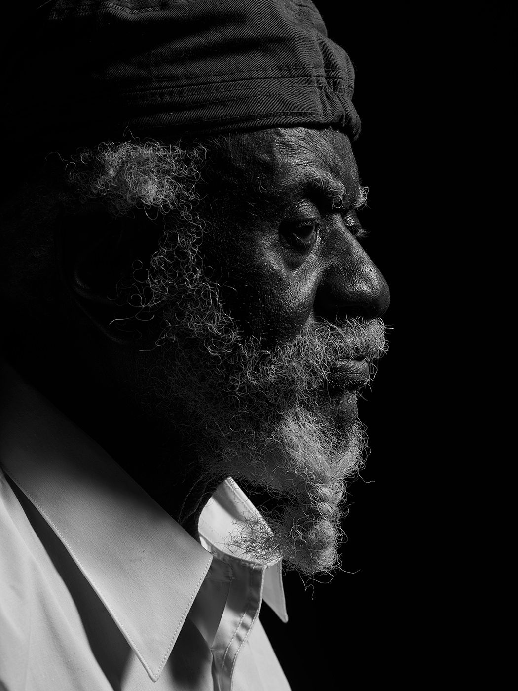 Black and white profile portrait of an elderly bearded man wearing a dark cap, dramatic chiaroscuro lighting. Pharoah Sanders, musician