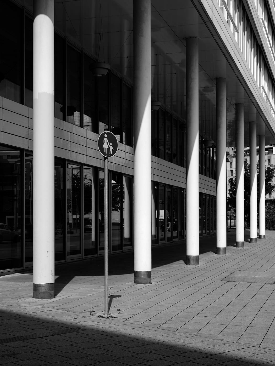Black and white photo of a modern building facade with white columns and a pedestrian sign on a sunny sidewalk.