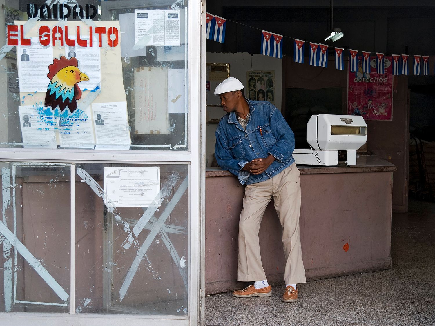 Man leaning against counter at El Gallito store in Cuba, Cuban flags decorating the ceiling above.