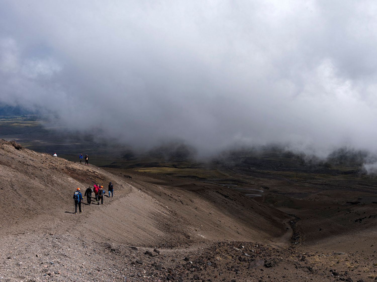 Hikers trek along a volcanic crater ridge under dramatic storm clouds with sweeping valley views.