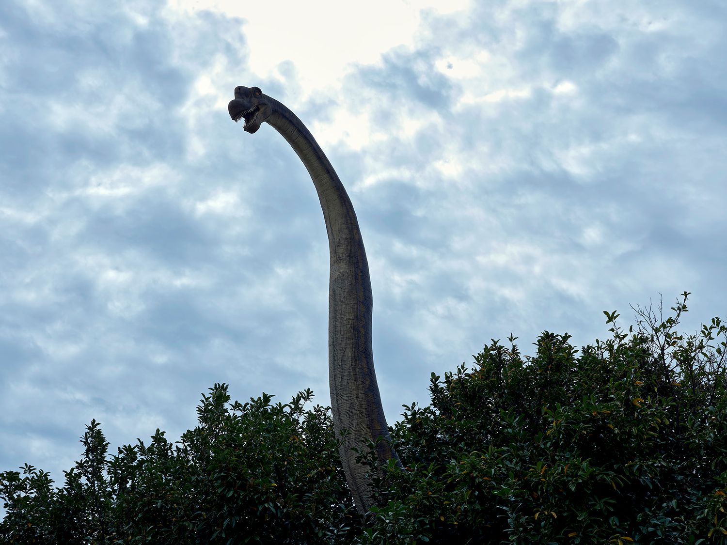 A towering dinosaur statue with a long neck rises above green trees against a cloudy sky.