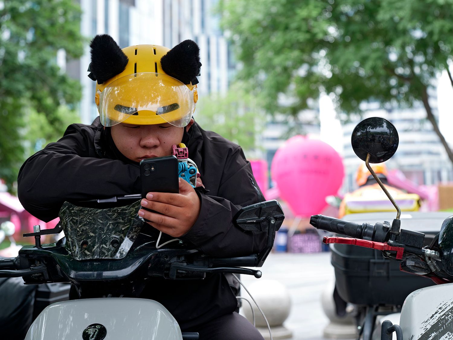 Person wearing a yellow cat ear helmet using a smartphone while sitting on a scooter outdoors.