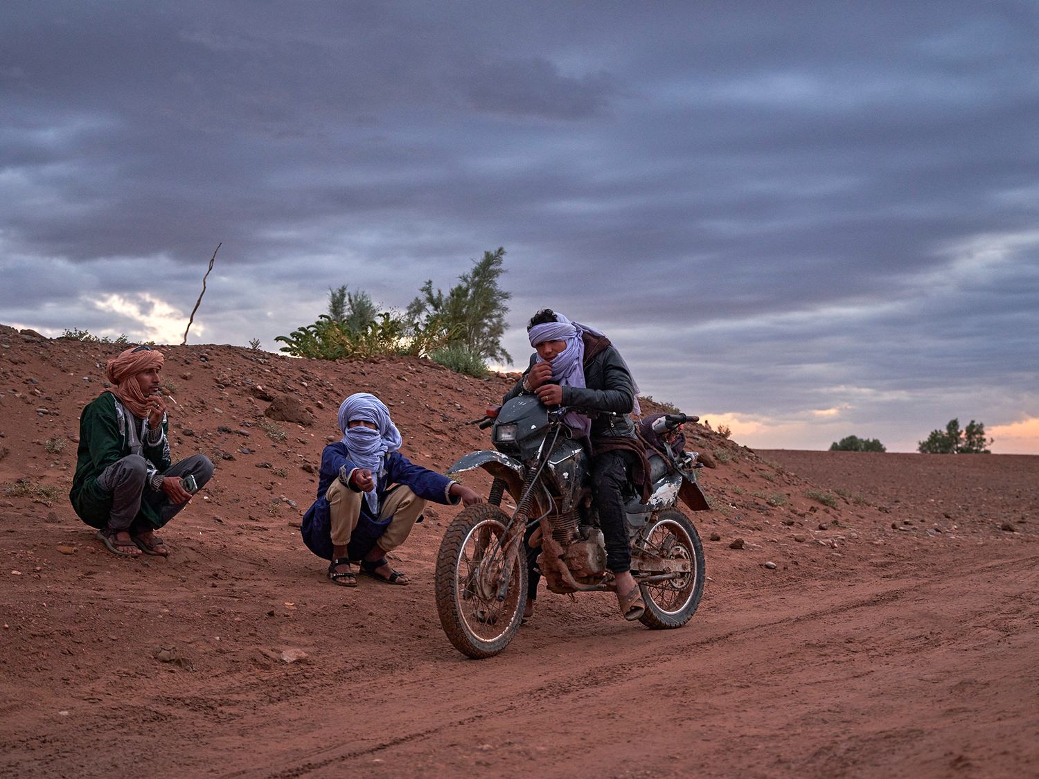 Men in traditional attire assist a dirt bike rider on a red sandy desert trail at dusk under a cloudy sky.