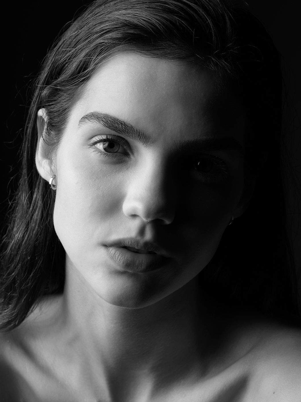 Black and white portrait of a young woman with dark hair, subtle earring, and intense gaze against dark background.