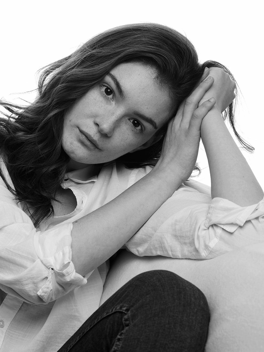 Black and white portrait of a young woman with dark wavy hair, wearing a white shirt and jeans, posing thoughtfully.