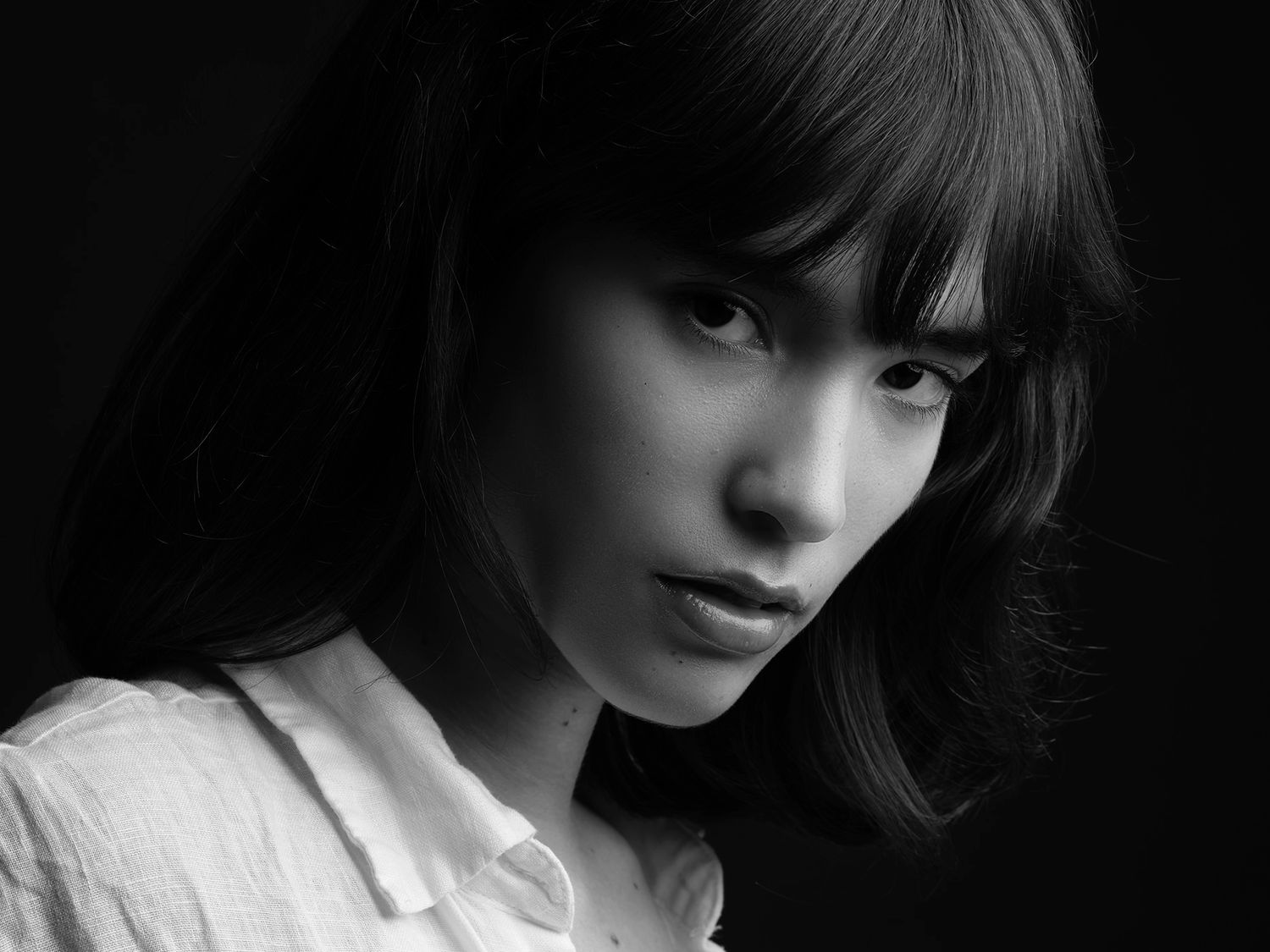 Black and white portrait of a young Asian woman with bangs, wearing a white shirt, against a dark background.