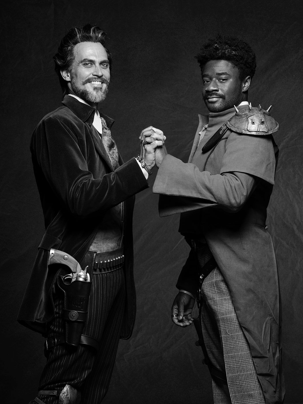 Two men in period costumes shake hands in a dramatic black and white portrait photo.