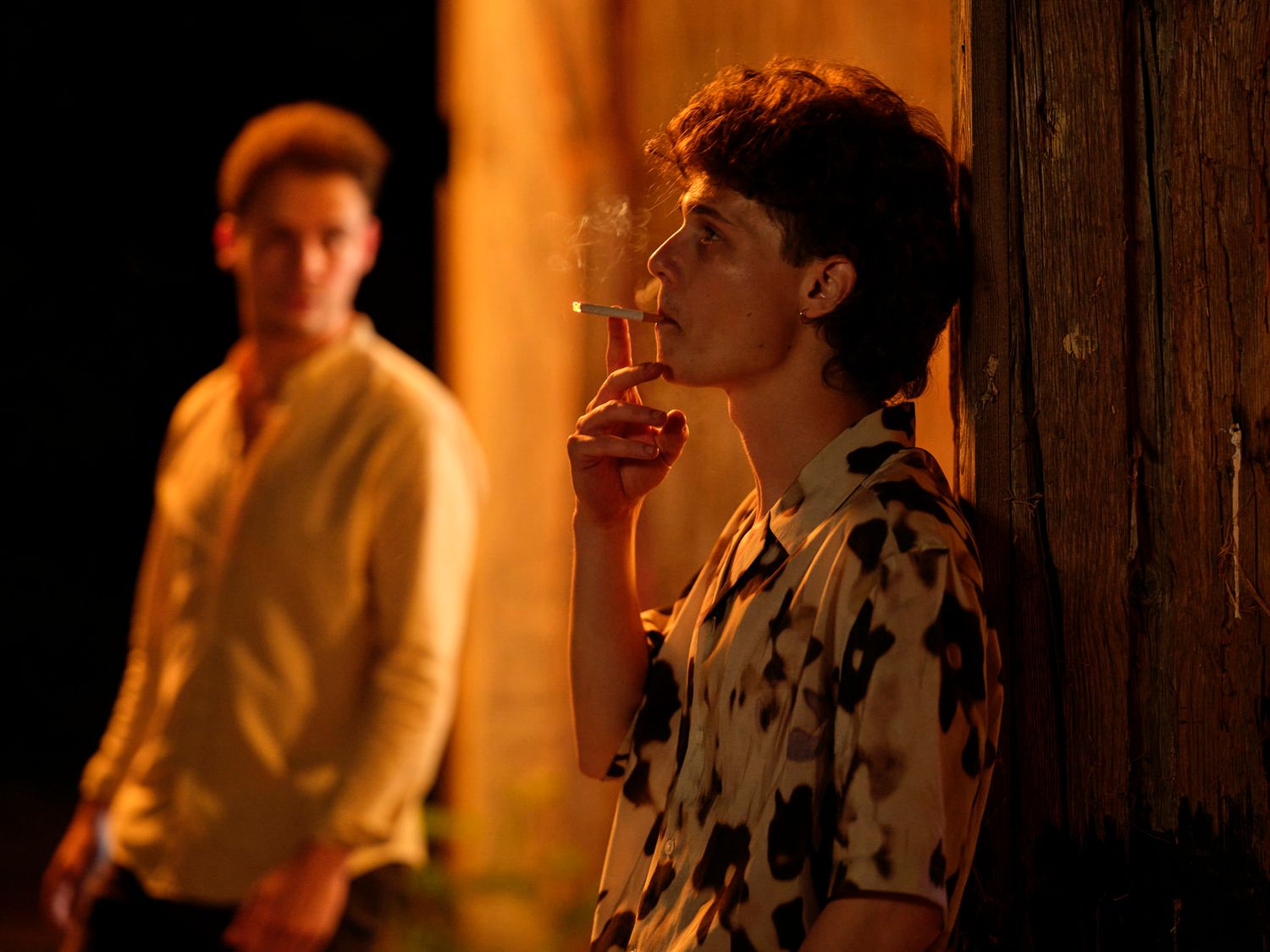 Two young men near a wooden wall, one smoking a cigarette in warm amber light.