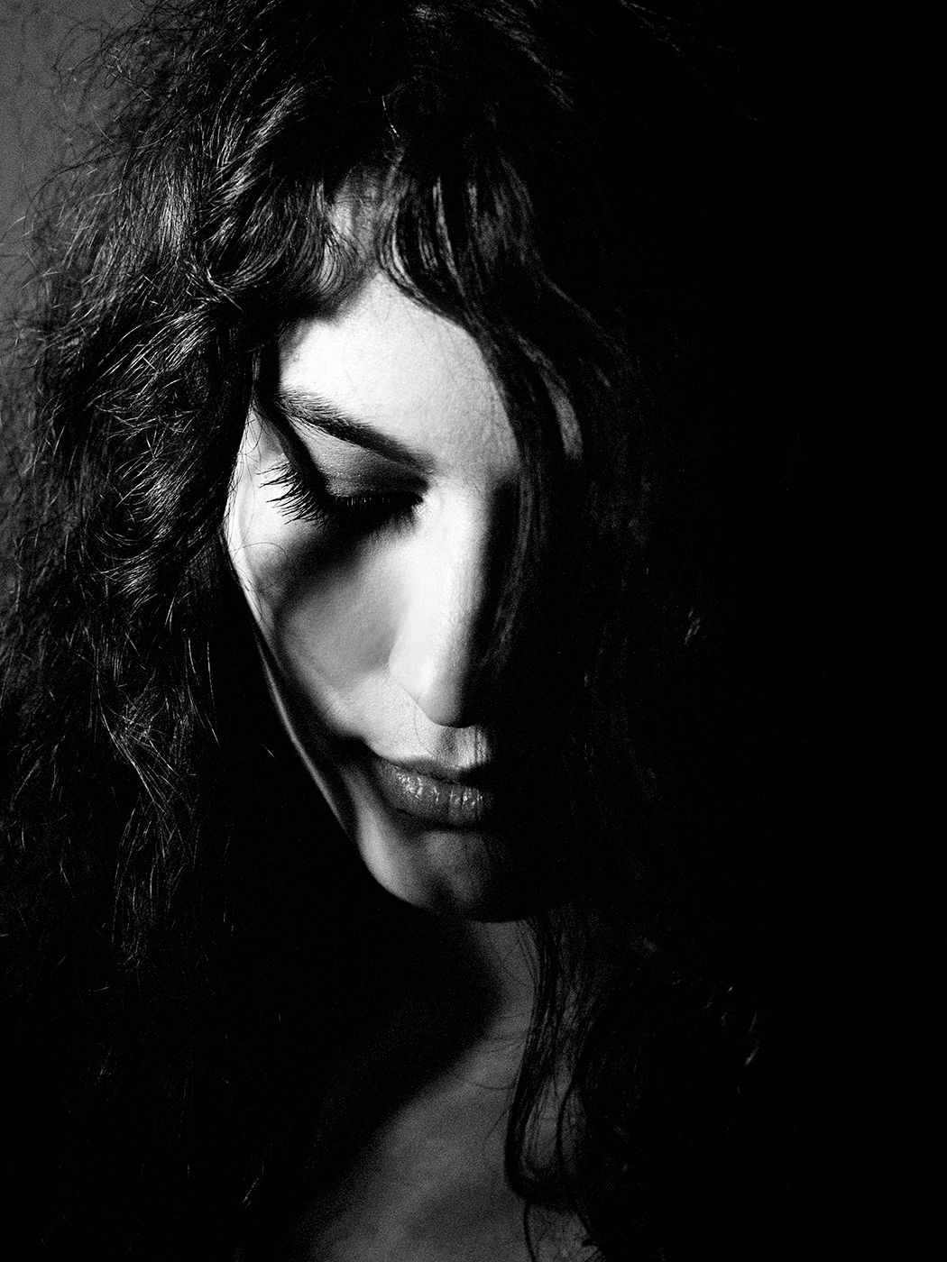 Black and white portrait of a woman with curly dark hair, eyes closed, head bowed in a dramatic low-key lighting setup.
