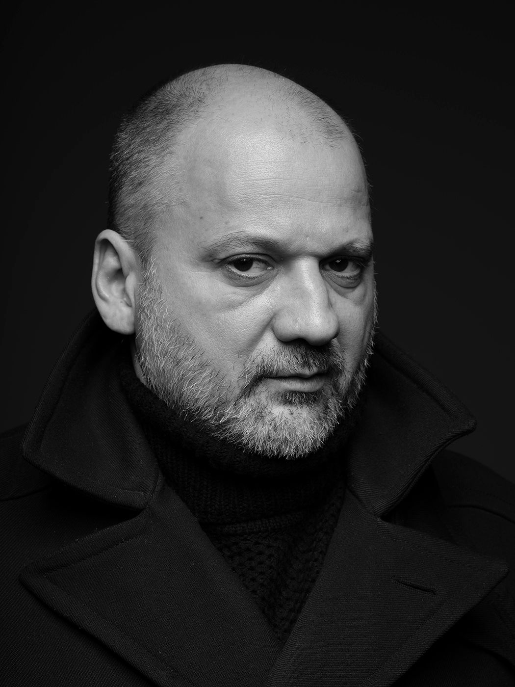 Black and white portrait of a bald man with a beard wearing a dark coat, serious expression.