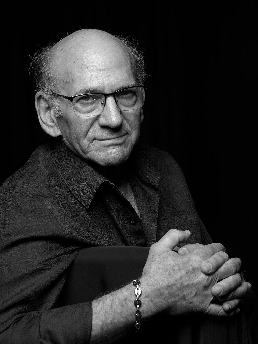 Black and white portrait of an elderly bald man with glasses, wearing a dark shirt, hands clasped, against dark background. Dave Liebman