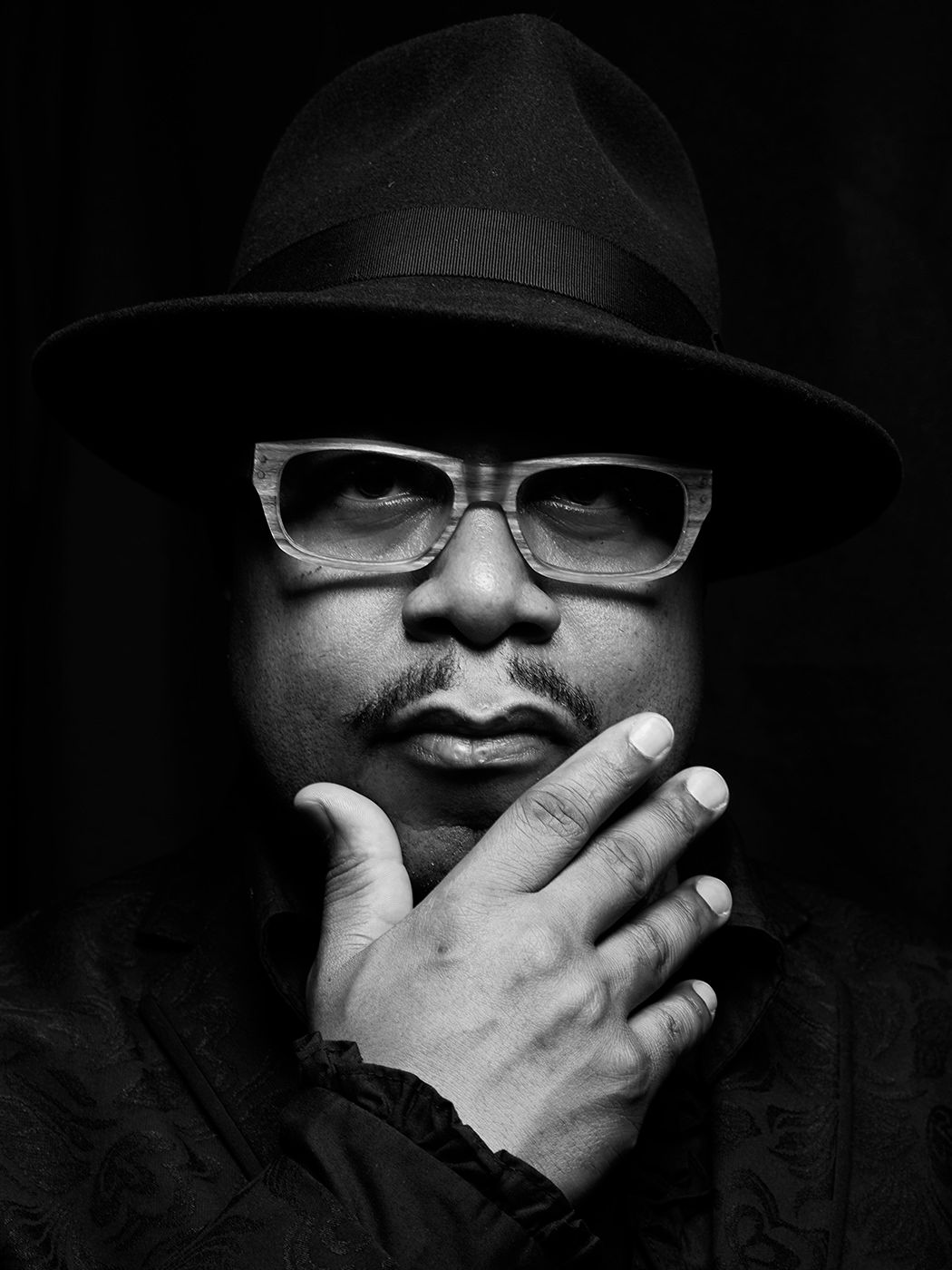 Black and white portrait of a man wearing a fedora hat and glasses, thoughtfully resting his hand on his chin. Nicholas Payton
