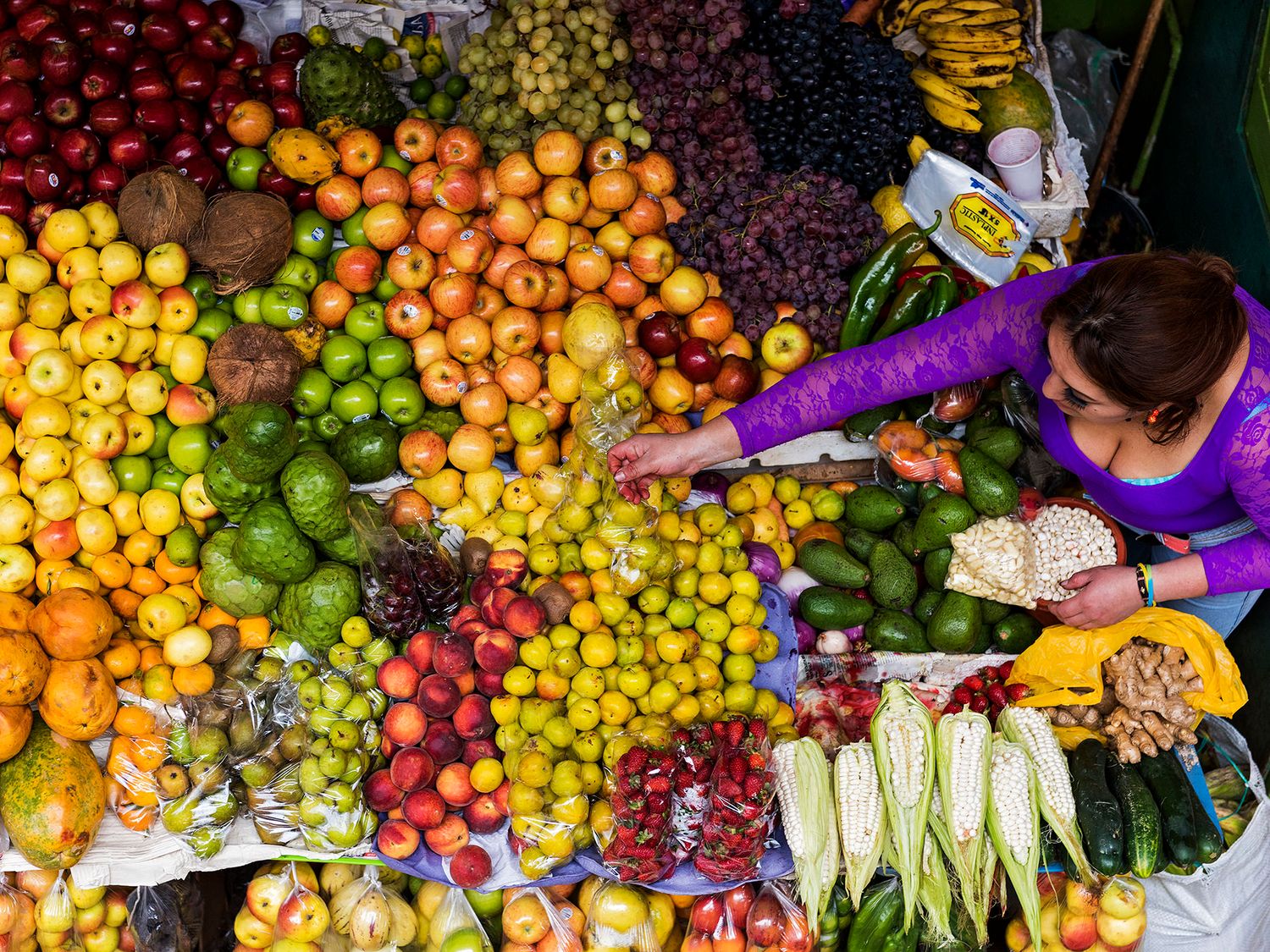Aerial view of a vibrant fruit market stall with colorful tropical fruits and a woman in purple selecting produce.
