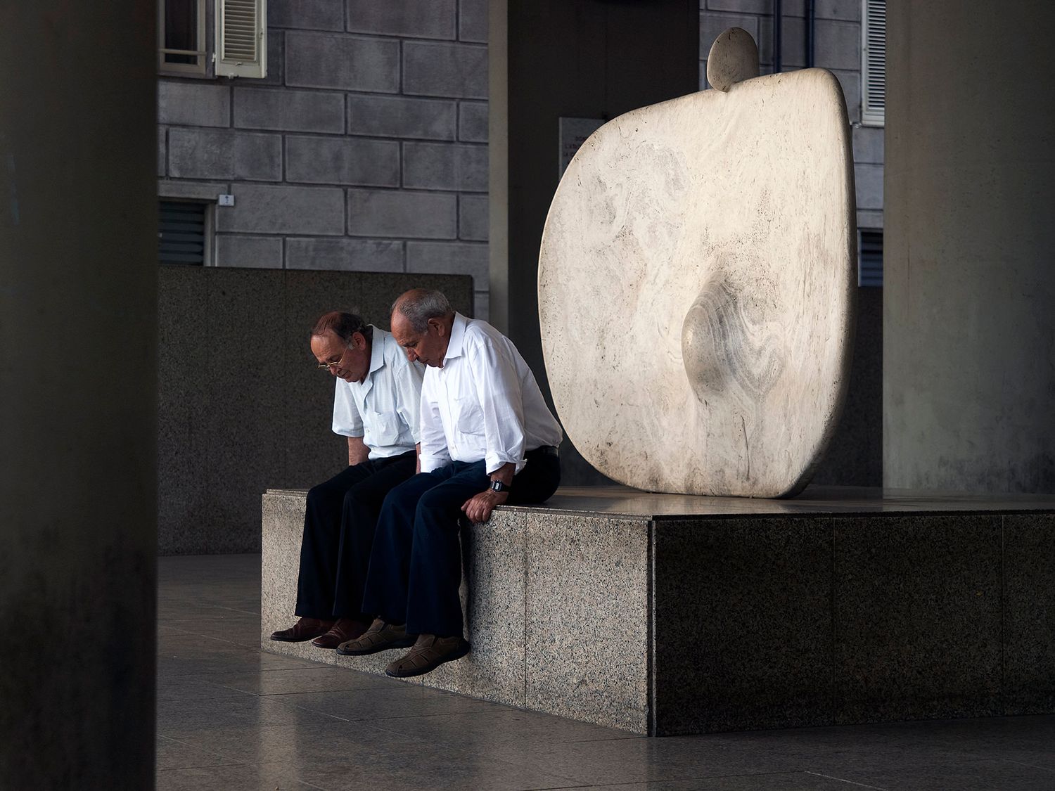Two elderly men in white shirts sit on a stone bench beside a large circular marble sculpture outdoors.